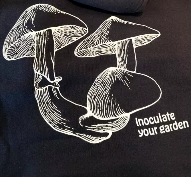 Navy blue hoodie with white line art: two mushrooms, small plant behind. Text: Inoculate Your Garden.